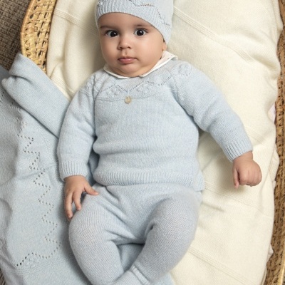 Bebé vestido com conjunto de malha azul clara de camisola, calças e gorro com pompom, deitado num cesto com manta creme.