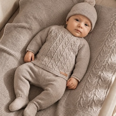 Bebé vestido com conjunto de malha bege claro deitado em manta de malha bege.