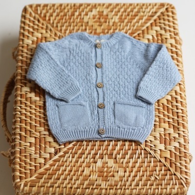 Casaco de malha azul bebé com botões de madeira sobre mala de vime trançado