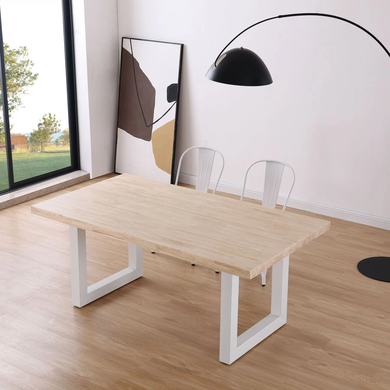Mesa de madeira clara com pernas brancas, cadeiras metálicas brancas, candeeiro de chão cinzento e quadro art abstrata ao fundo