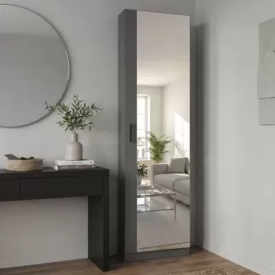 Armário alto cinzento com porta espelhada ao lado de secretária preta com vaso e livros, numa sala com chão de madeira e tapete bege.