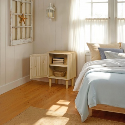 Mesa de cabeceira em madeira clara com porta aberta ao lado de cama com roupa azul clara.