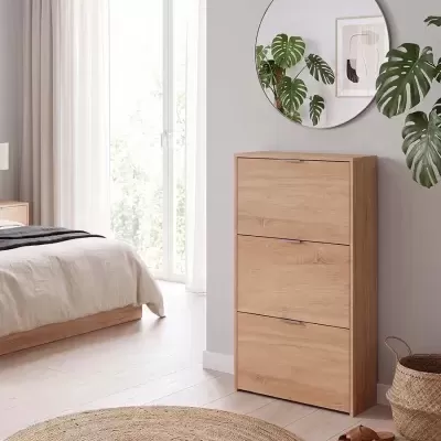 Móvel de madeira clara com três gavetas junto a uma cama branca, tapete redondo e espelho redondo na parede