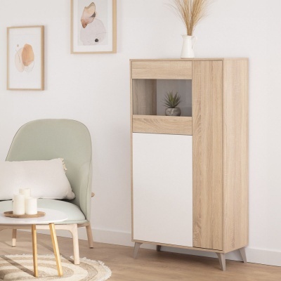 Móvel de madeira clara com porta branca e compartimento de vidro, cadeira verde claro com almofada, mesa com velas, quadros e vaso com plantas secas.