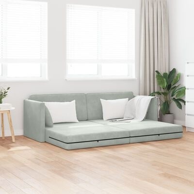 Sofá-cama cinzento claro com almofadas brancas numa sala luminosa com chão de madeira e plantas.
