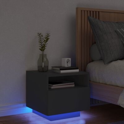 Mesa de cabeceira preta com luz LED azul na base ao lado de cama com cabeceira de madeira e almofadas cinzentas