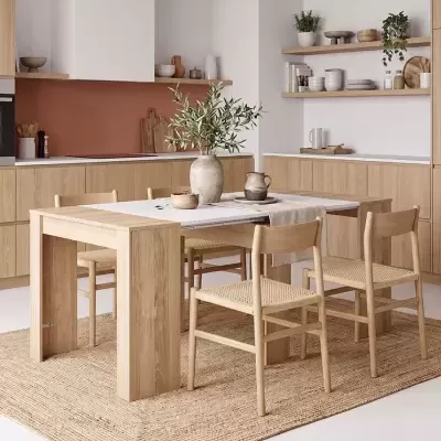 Mesa de jantar extensível de madeira clara com cadeiras de madeira e assentos em fibra natural num ambiente de cozinha moderna.