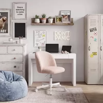 Escritório em casa com secretária branca, cadeira rosa, puff azul, armário com stickers e decoração de parede com quadros e plantas