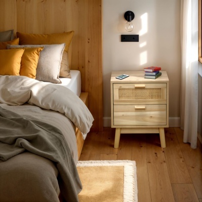Mesa de cabeceira em madeira clara com gavetas, cama com almofadas mostarda e bege em quarto iluminado