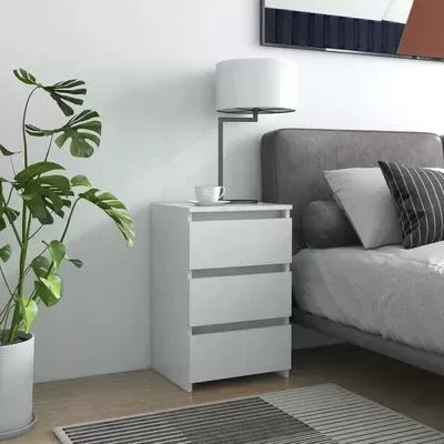 Móvel branco com três gavetas, candeeiro e chávena, cama cinza com almofadas, planta e tapete