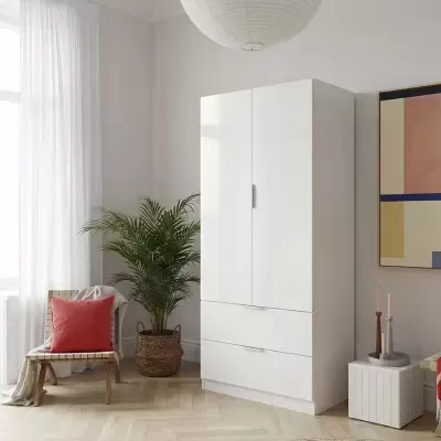 Armário branco de madeira com duas portas e gavetas numa sala, cadeira com almofada vermelha e planta em vaso ao lado