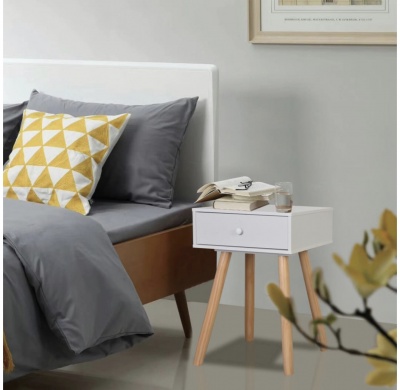 Cama cinzenta com almofada geométrica amarelo e branca ao lado de mesa de cabeceira branca com pernas de madeira