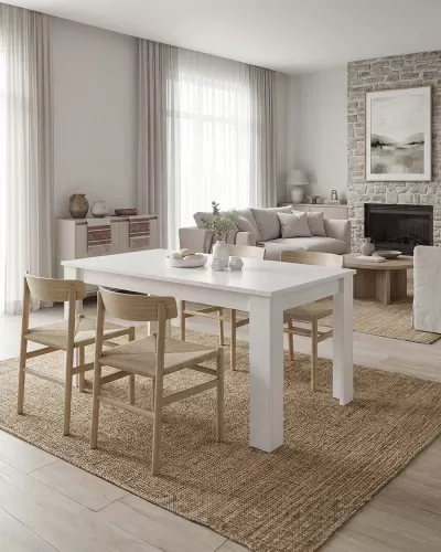 Sala de jantar com mesa branca e quatro cadeiras de madeira clara sobre tapete castanho claro, sofá bege e lareira em fundo de pedra.