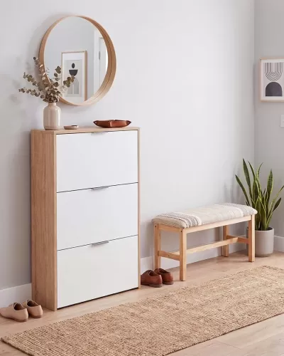 Móvel para sapatos branco e madeira com espelho redondo, banco estofado e planta num ambiente interior moderno