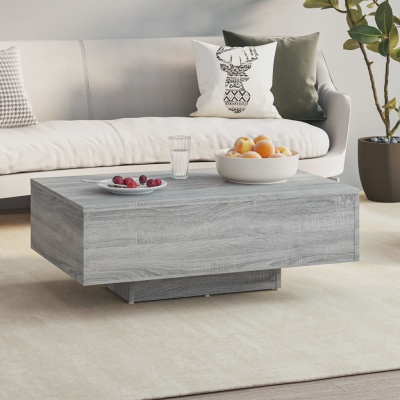 Mesa de centro cinza claro rectangular com superfície de madeira, decorada com frutas e copo, junto a sofá creme com almofadas e planta em vaso cinza.
