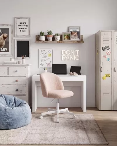Escritório em casa com secretária branca, cadeira rosa, puff azul, armário com stickers e decoração de parede com quadros e plantas