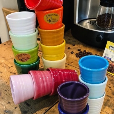 Vários copos pequenos coloridos em cerâmica com etiquetas douradas, numa mesa de madeira com máquina de café e grãos de café.