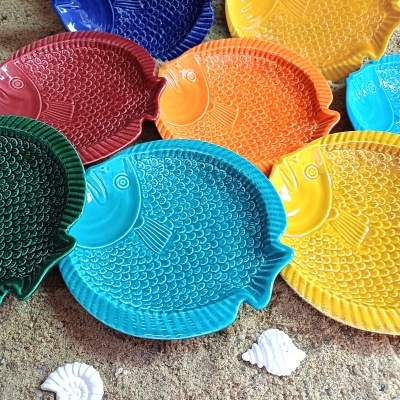 Pratos coloridos em forma de peixe com textura de escamas sobre areia com conchas.