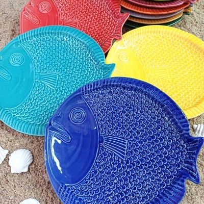 Pratos coloridos em cerâmica em formato de peixe com textura de escamas e conchas numa superfície de areia