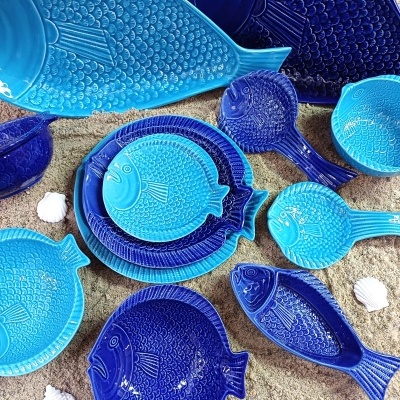 Conjunto de loiça em cerâmica com pratos e taças azuis em forma de peixe sobre fundo imitando areia.