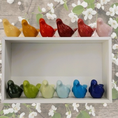 Pássaros decorativos de cerâmica colorida organizados em prateleira branca sobre fundo floral.