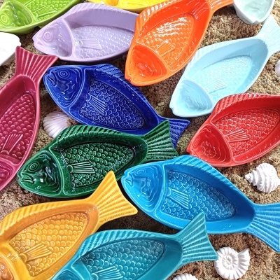 Pratos cerâmicos coloridos com formato de peixe sobre tecido castanho com conchas brancas