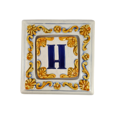Azulejo cerâmico decorativo com letra H azul e padrão floral amarelo e azul.