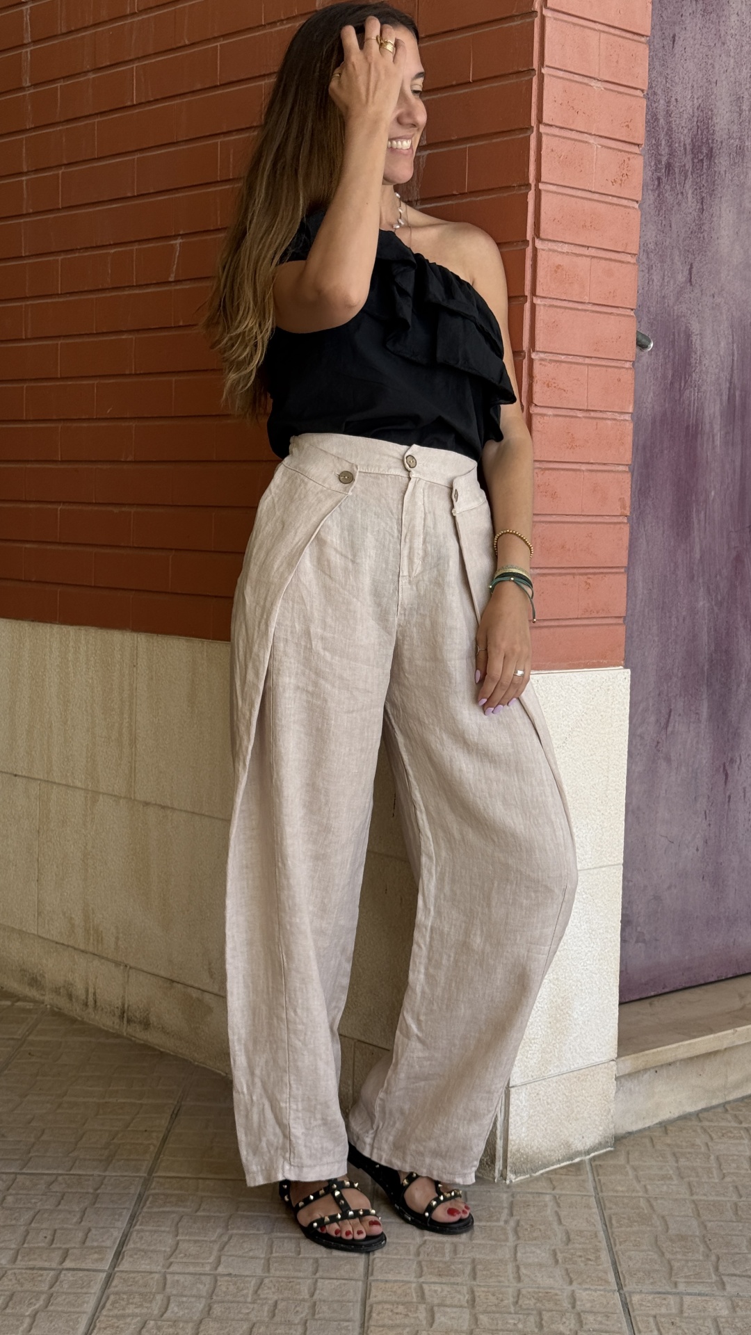 Mulher com calças de linho bege largas e blusa preta de um ombro, sandálias pretas, parede de tijolo vermelho e chão cinzento