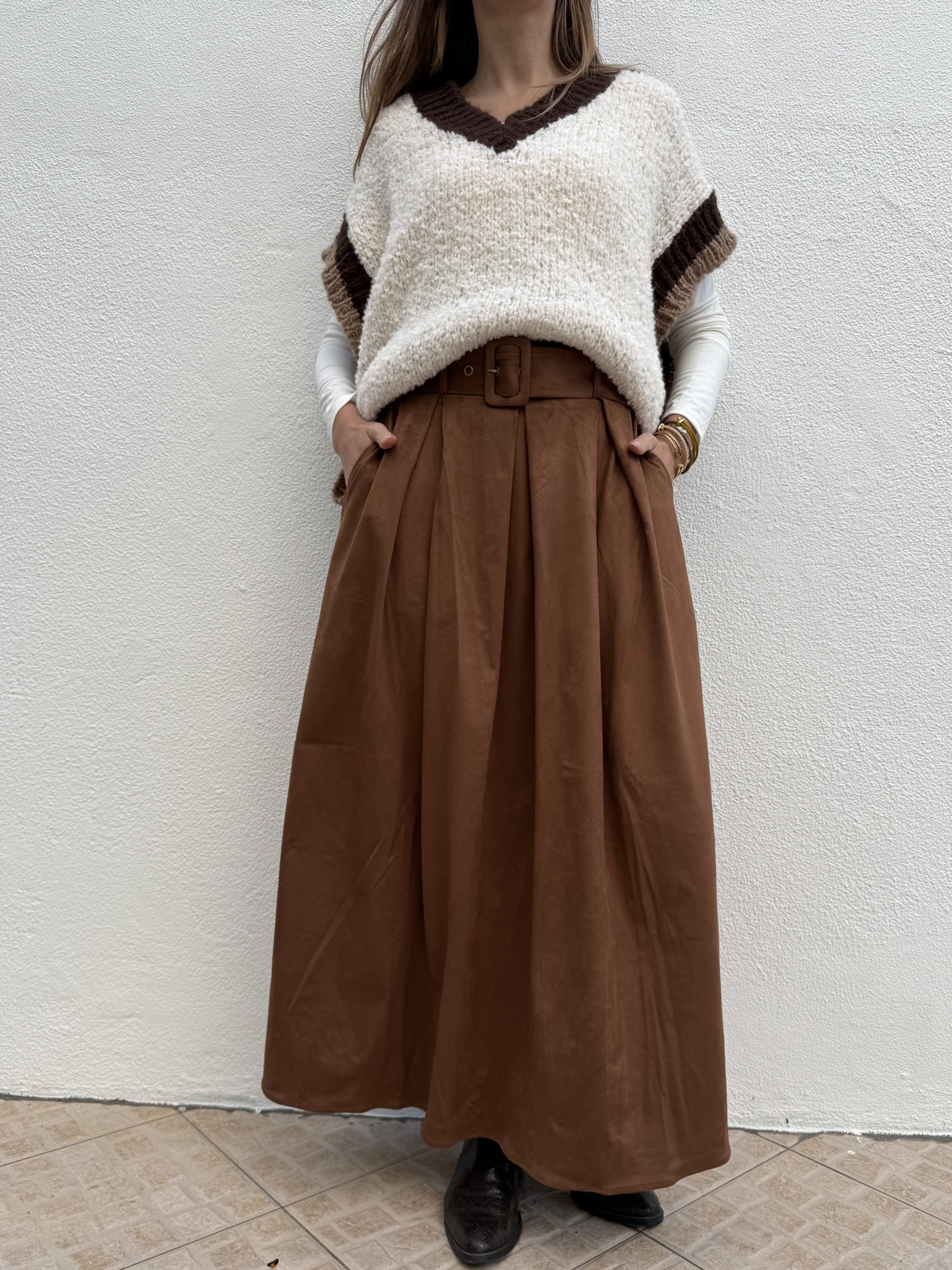 Mulher com saia comprida castanha e camisola de malha branca e castanha em fundo de parede branca.