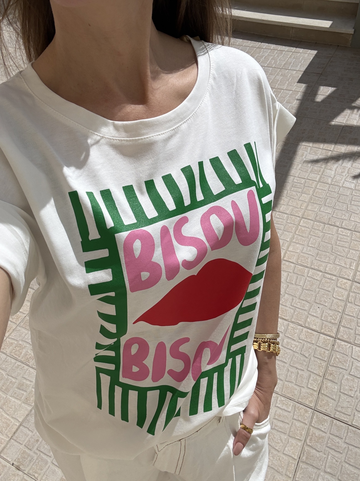 T-shirt branca com estampado verde, rosa e vermelho com texto BISOU BISOU, usada com calças brancas e joias douradas