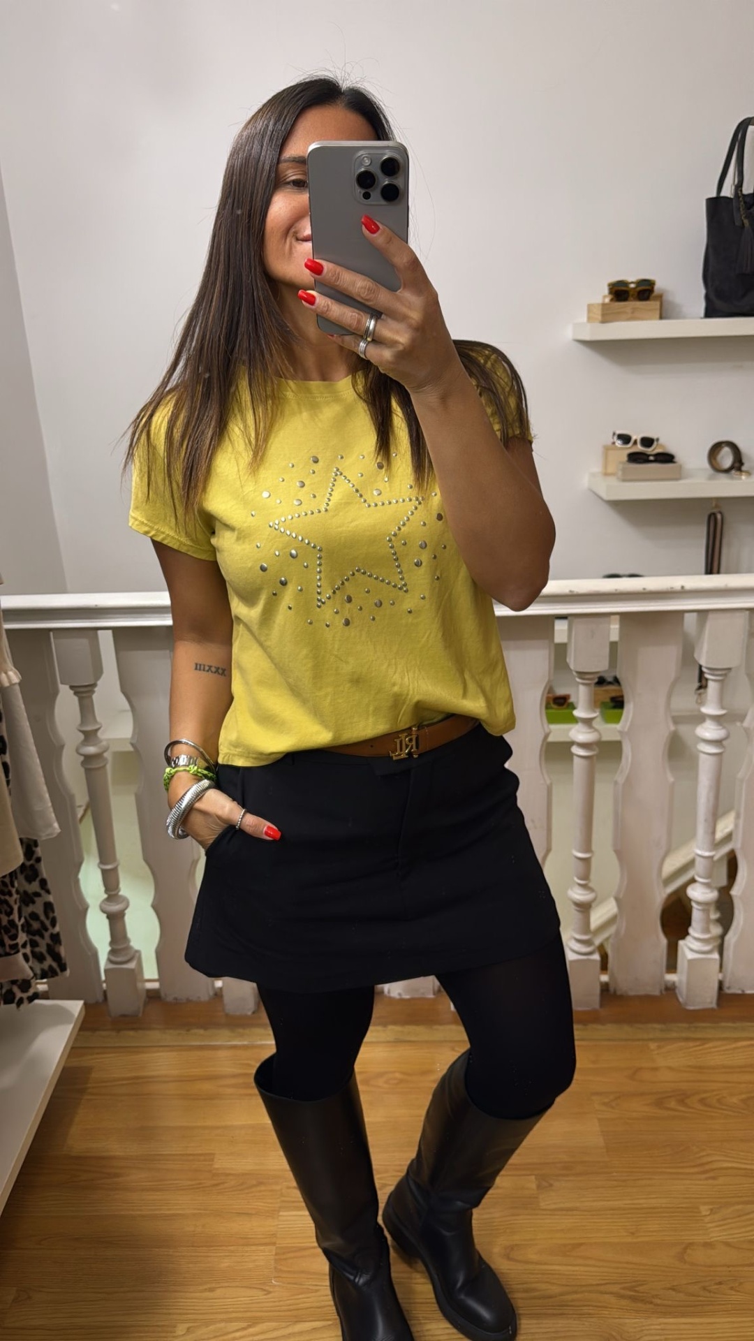 Mulher vestindo camisola amarela com estrela de tachas, saia preta, botas pretas em interior com chão de madeira