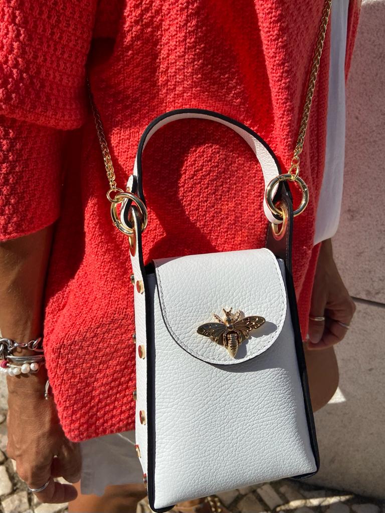 Bolsa branca em pele texturada com abelha dourada, alça de corrente dourada, com casaco vermelho ao fundo