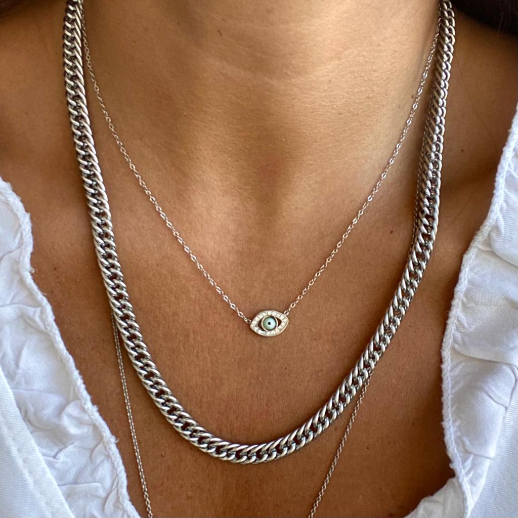 Dois colares prateados, um grosso e outro fino com pendente de olho azul, usado com camisa branca