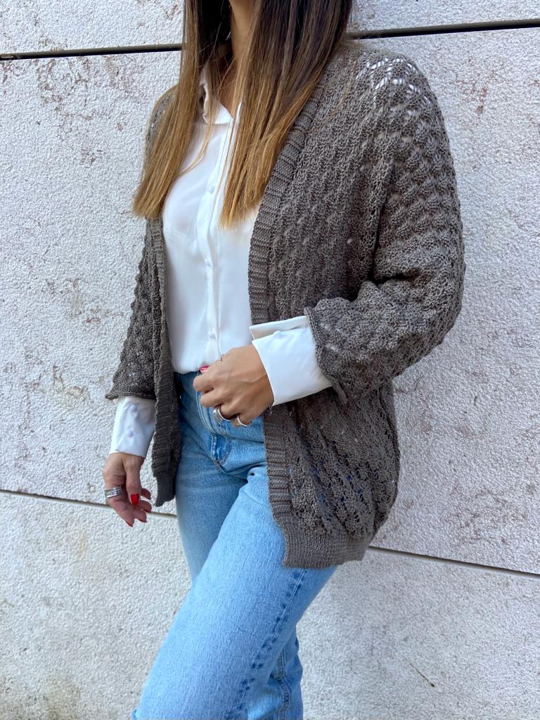 Mulher com camisola branca, casaco cinza de malha e calças de ganga azuis junto a uma parede de pedra.