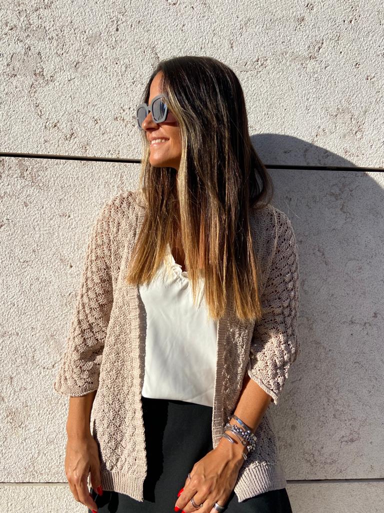 Mulher com cardigan bege tricotado, camisa branca e calças pretas junto a parede de pedra clara