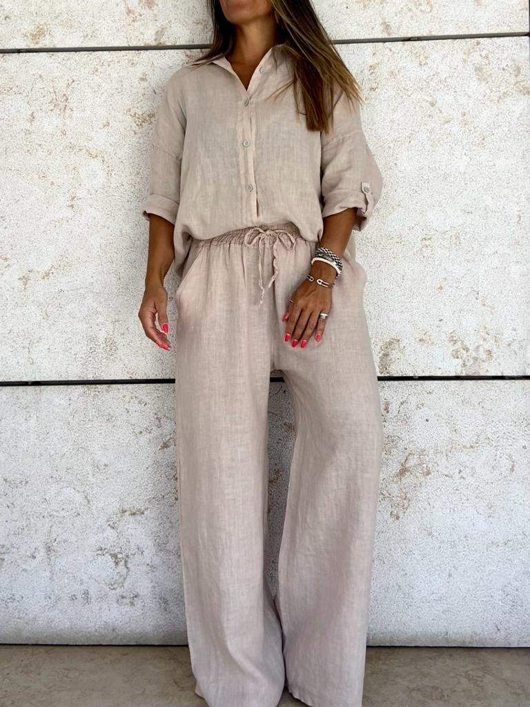 Conjunto feminino bege claro com camisa de manga comprida e calças largas, em fundo de parede de pedra clara