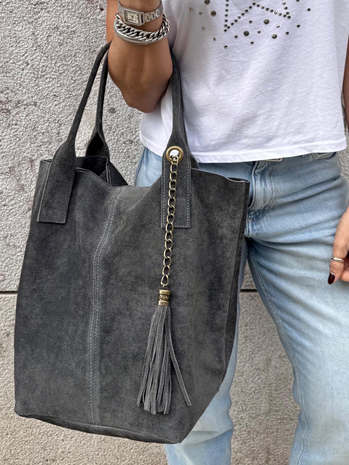 Mala de mão cinzenta em pele camurça com tassel, alças duplas, junto a pessoa com calças de ganga e t-shirt branca.