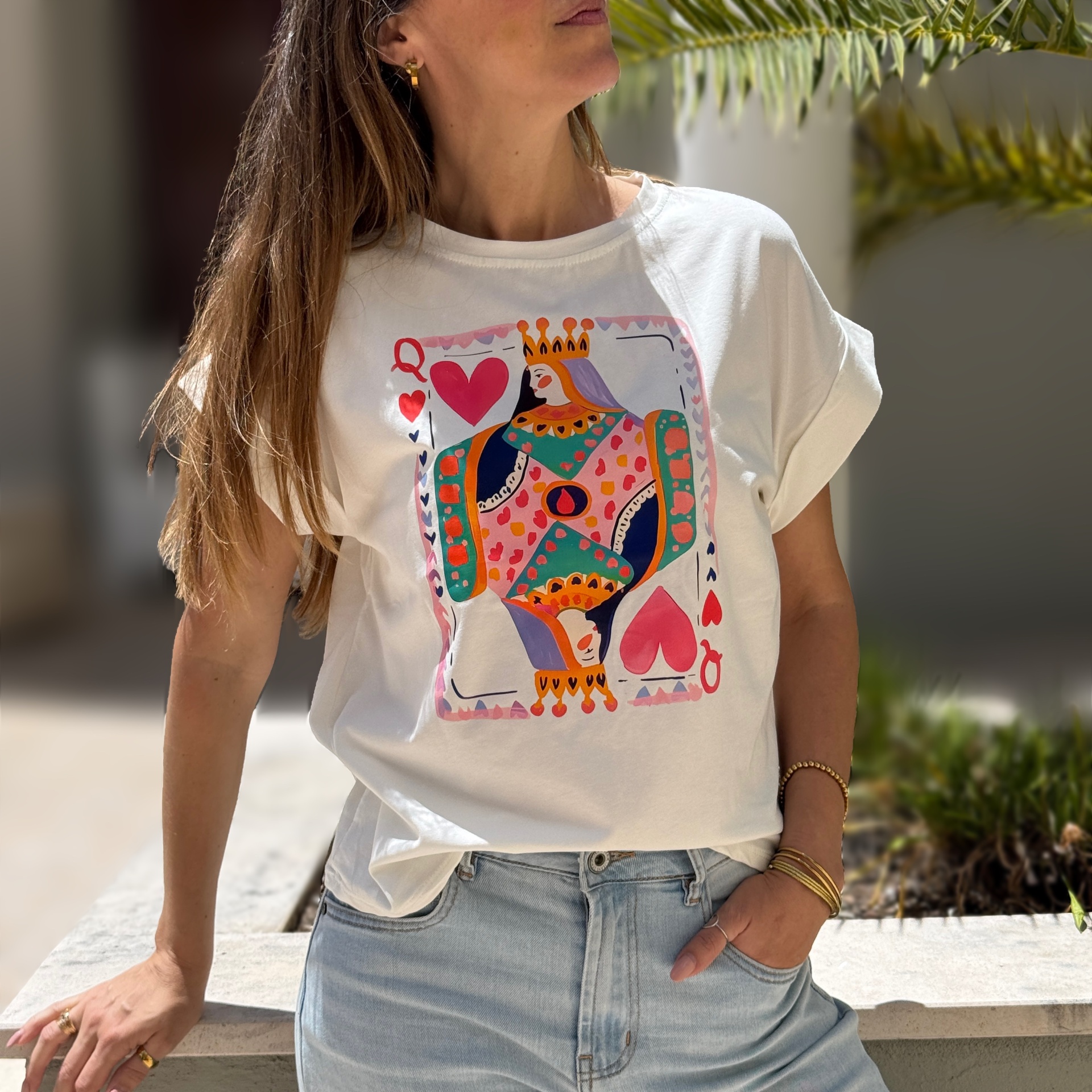 Mulher a usar t-shirt branca com estampado colorido da Rainha de Copas e calças de ganga claras, em ambiente exterior com plantas.