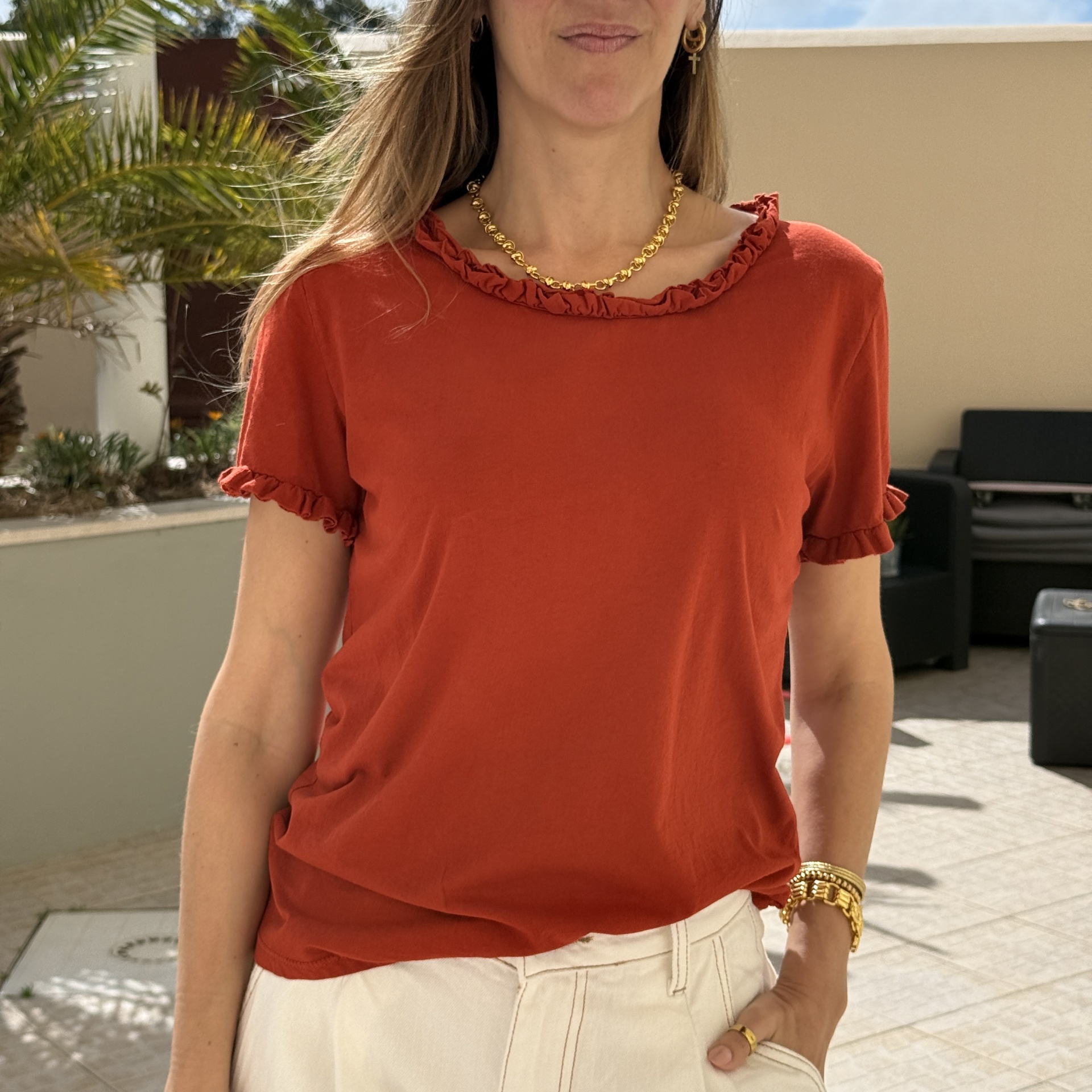 Mulher vestindo t-shirt vermelha-alaranjada com bordado franzido na gola e nas mangas, acessórios dourados, ao ar livre.