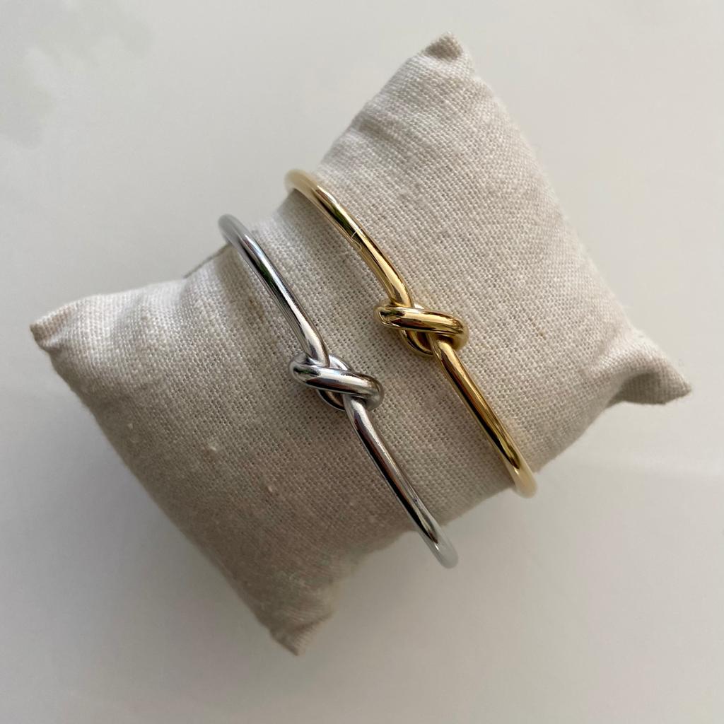 Duas pulseiras metálicas finas, prateada e dourada, com nó decorativo em almofada bege
