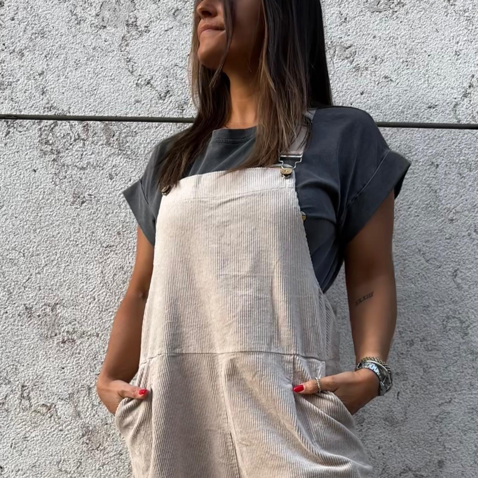 Pessoa vestindo macacão de tecido canelado cinza claro com alças ajustáveis e bolsos frontais, t-shirt preta, parede clara com textura