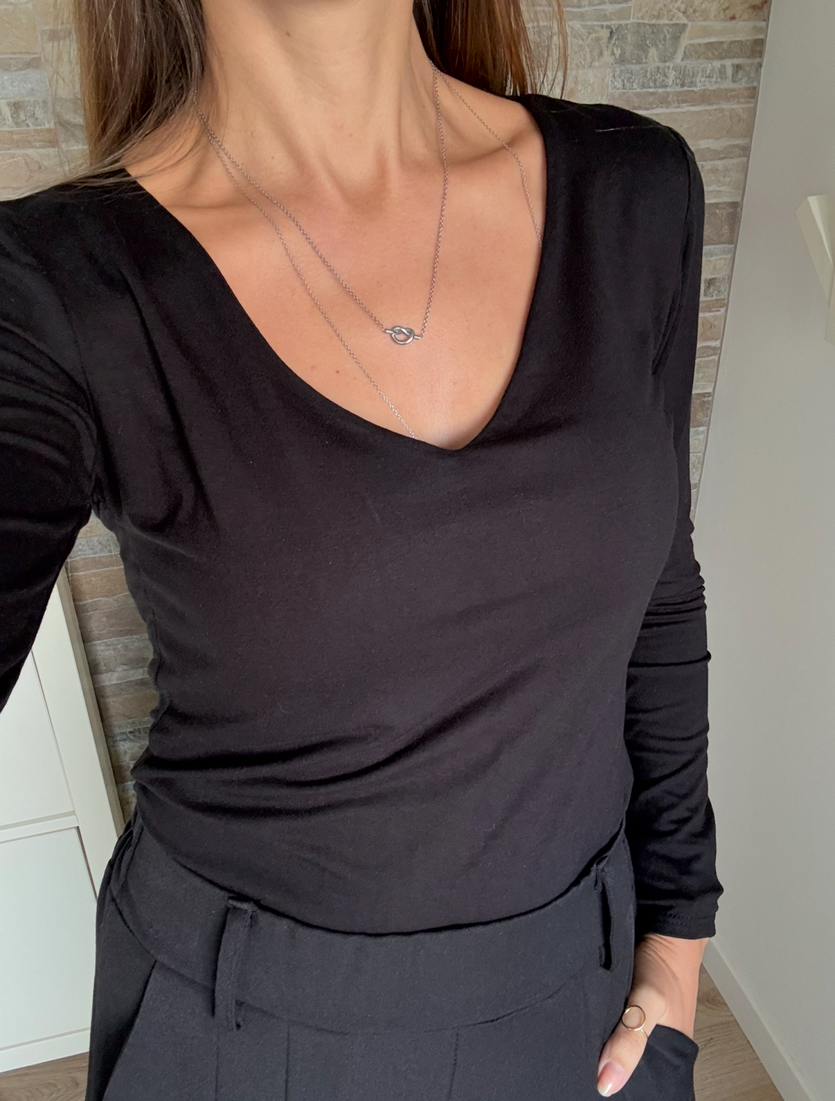 Pessoa usando camisa preta de manga comprida com decote em V, calças pretas e colar prateado com fundo interior