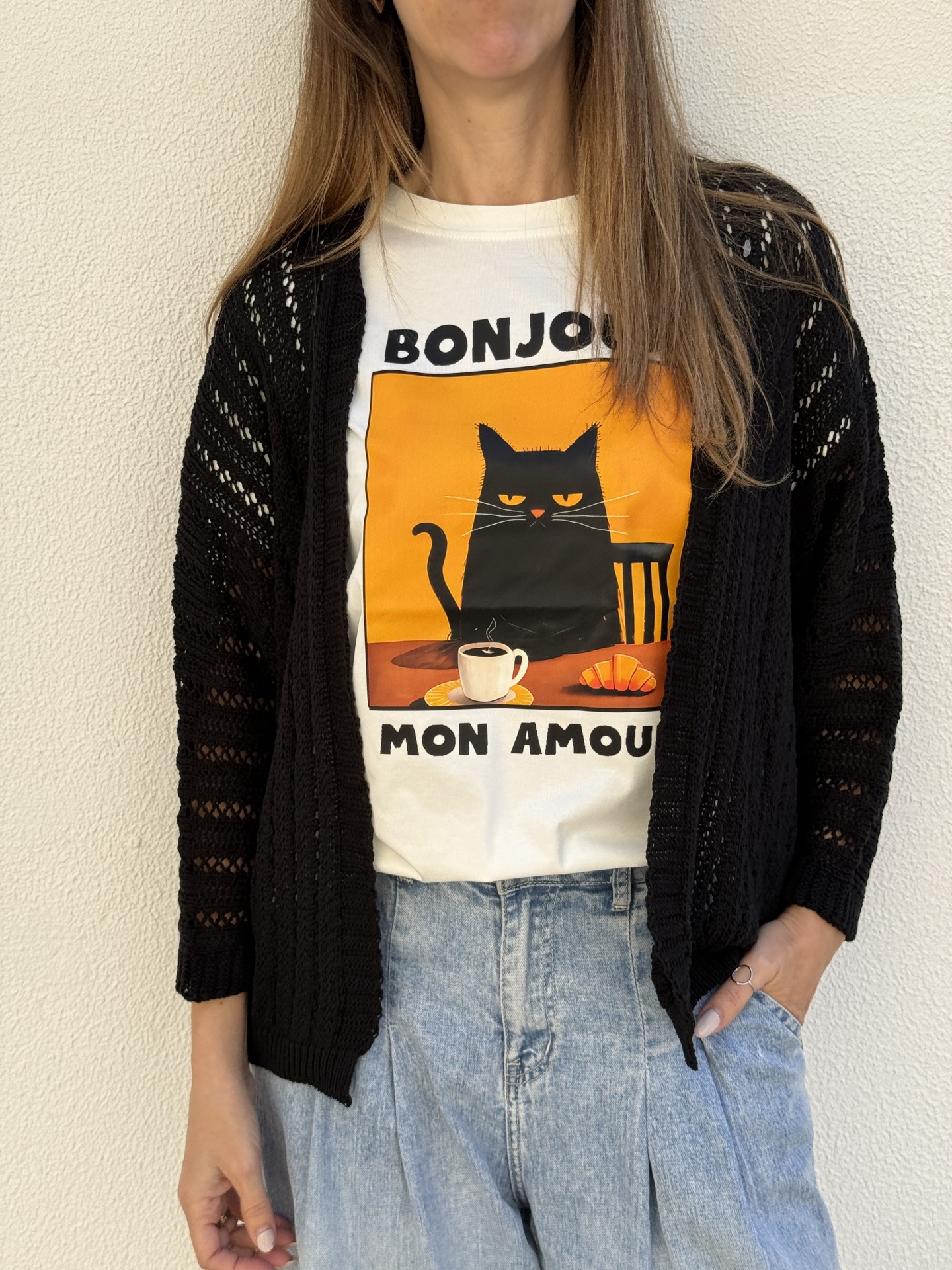 T-shirt branca com estampa de gato preto e texto BONJOUR MON AMOUR usada com casaco preto e calças de ganga azuis claras.