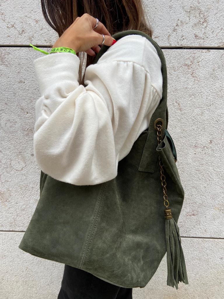 Pessoa segurando mala de camurça verde escura com franjas, fundo parede clara