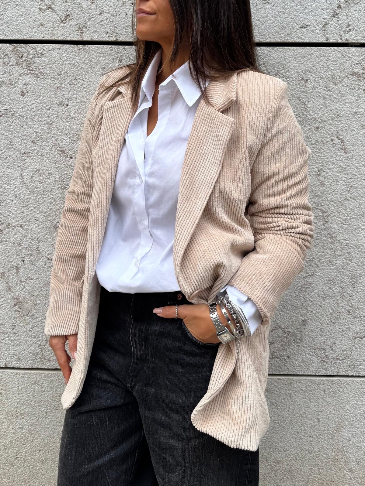 Pessoa com casaco bege de veludo cotelê, camisa branca e calças pretas de pé frente a parede cinza
