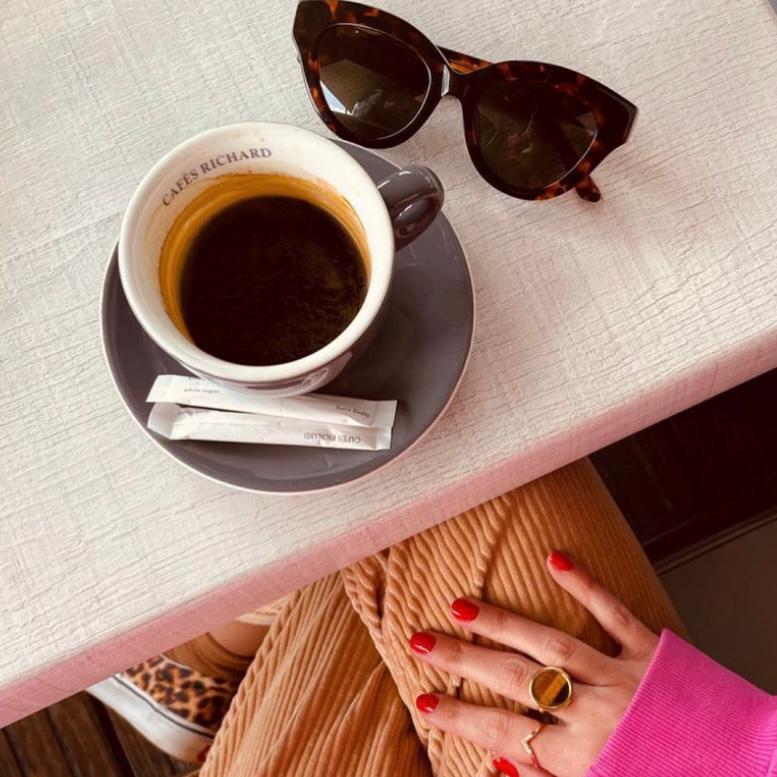 Chávena de café preto com pires cinzento, óculos de sol de padrão tartaruga e mão feminina com unhas vermelhas e anéis dourados