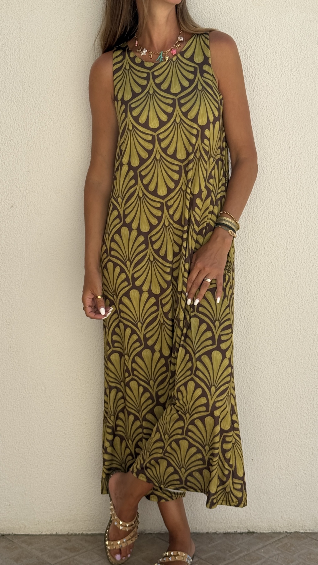 Vestido longo castanho com padrão verde de folhas largas, sem mangas, vestido por mulher com sandálias douradas com tachas.