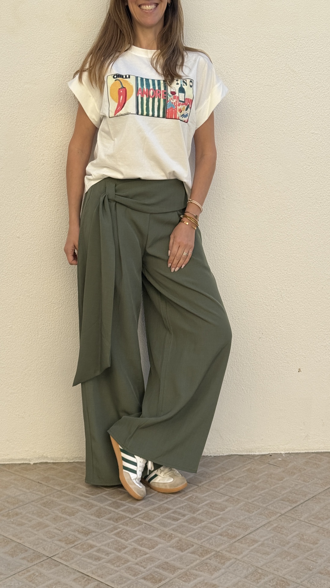 Mulher com t-shirt branca estampada e calças largas verdes com cinto e ténis brancos em chão de mosaico cinzento.