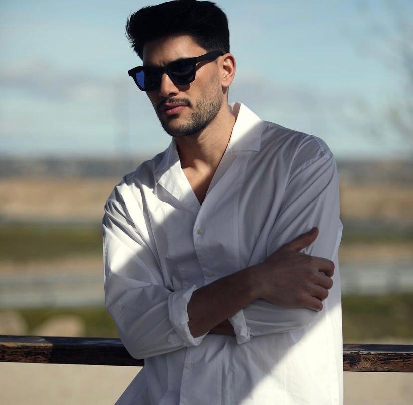 Homem de camisa branca e óculos de sol encostado em estrutura de madeira ao ar livre