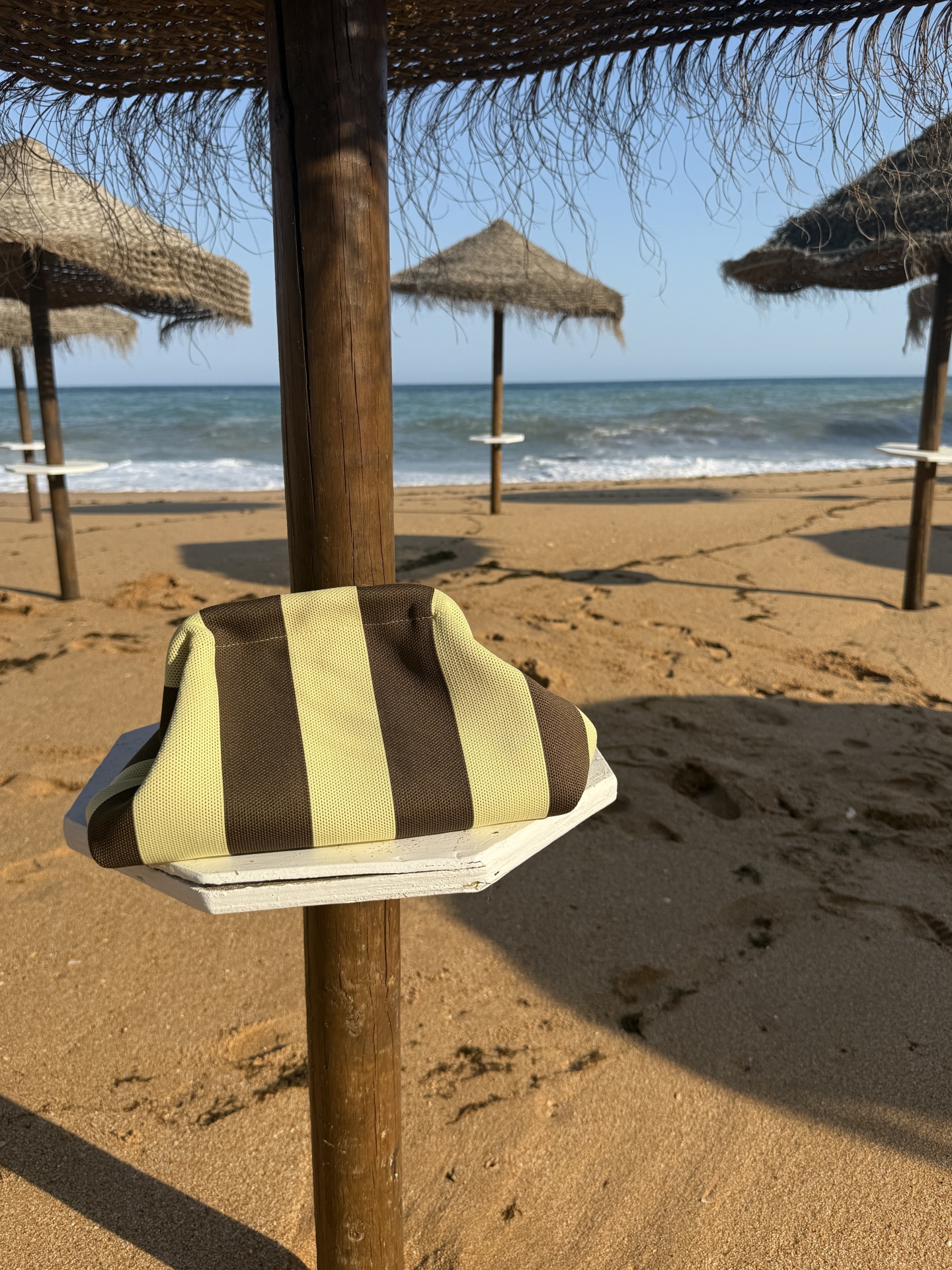 Guarda-sol de palha na praia com toalha às riscas castanhas e amarelas, mar ao fundo e areia dourada.